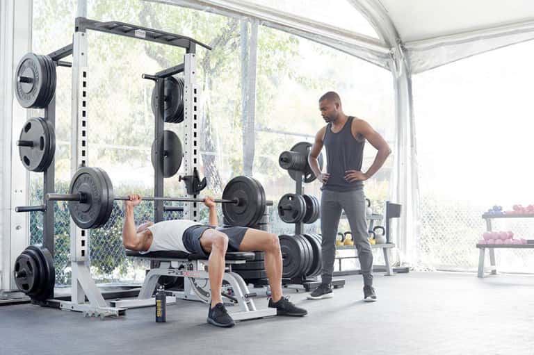 Personal trainer supervising man doing barbell bench press in modern gym with glass walls