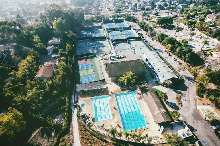 Aerial view of tennis courts and two blue swimming pools surrounded by lush green landscape