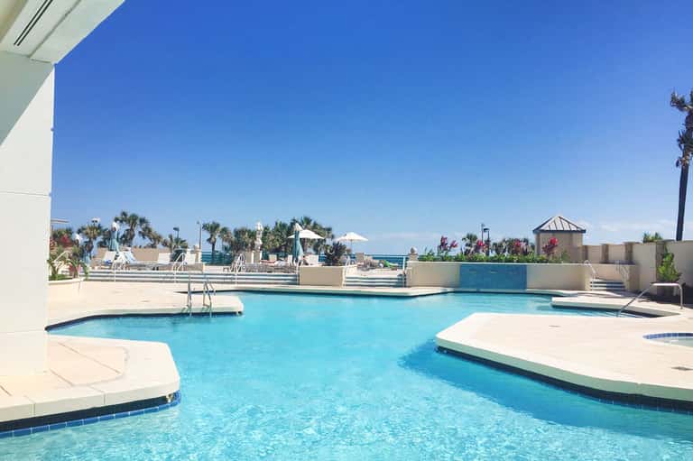 Outdoor resort pool with geometric design, palm trees, lounge areas, and ocean horizon view
