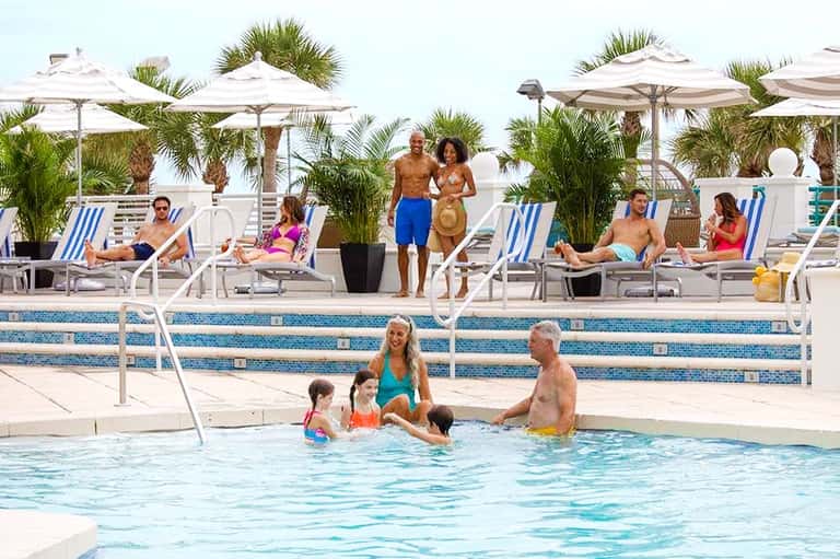 Resort pool area with families swimming, lounge chairs, umbrellas, and palm trees in background