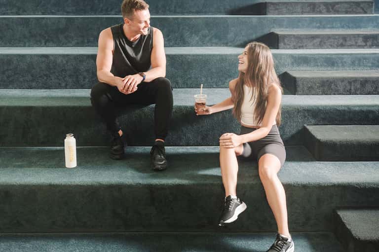 Two people in athletic wear sitting on green steps with water bottles, conversing and smiling