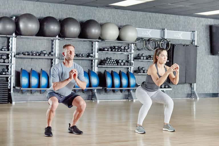 Couple performing squats in gym with exercise balls and dumbbells on shelving behind them