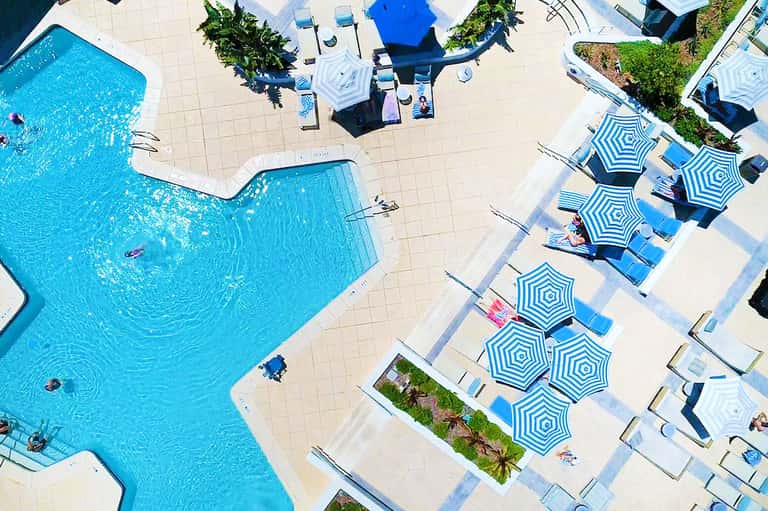 Aerial view of resort pool with lounge chairs, blue umbrellas, and surrounding landscaping