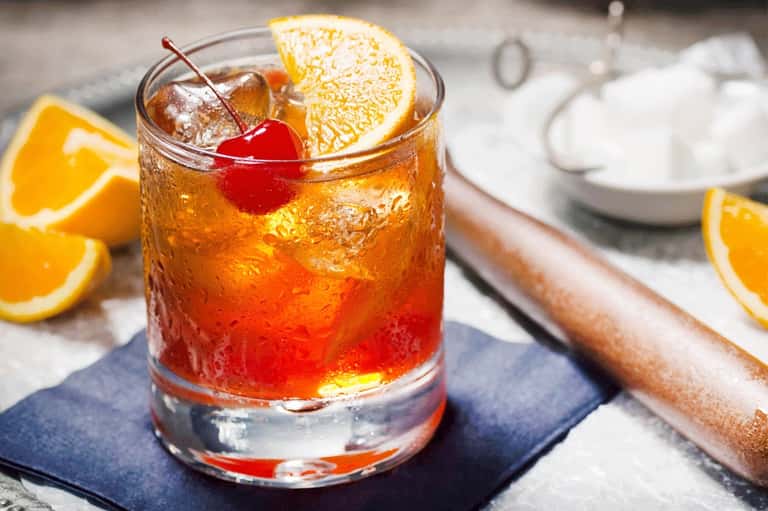 Cocktail with orange slice and cherry garnish on dark coaster with citrus slices nearby