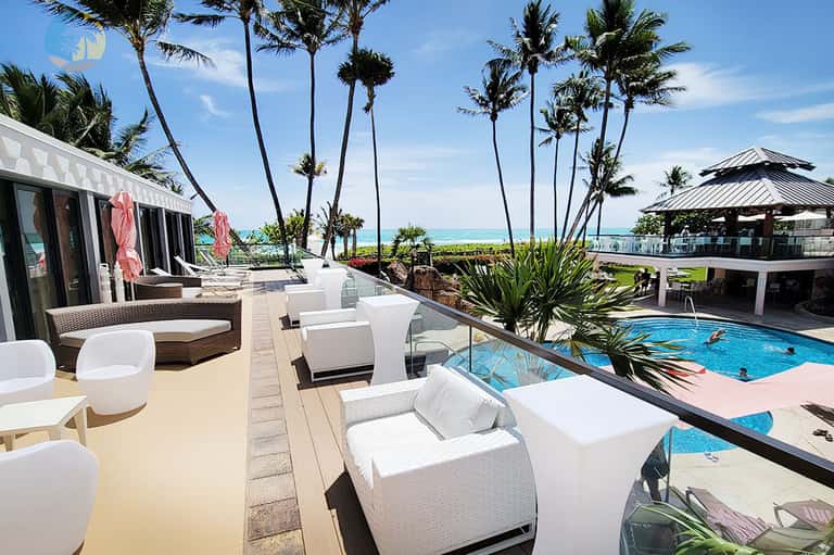 Beachfront resort pool deck with white lounge chairs, palm trees, ocean view, and modern architecture