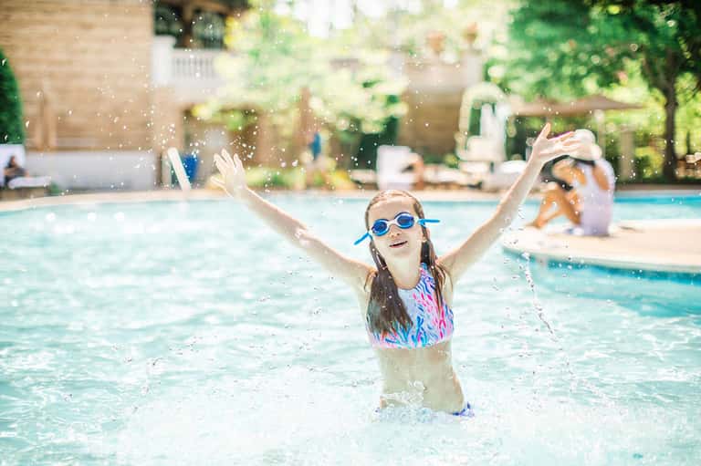 Child in pool wearing goggles with arms raised, splashing water in sunny resort setting