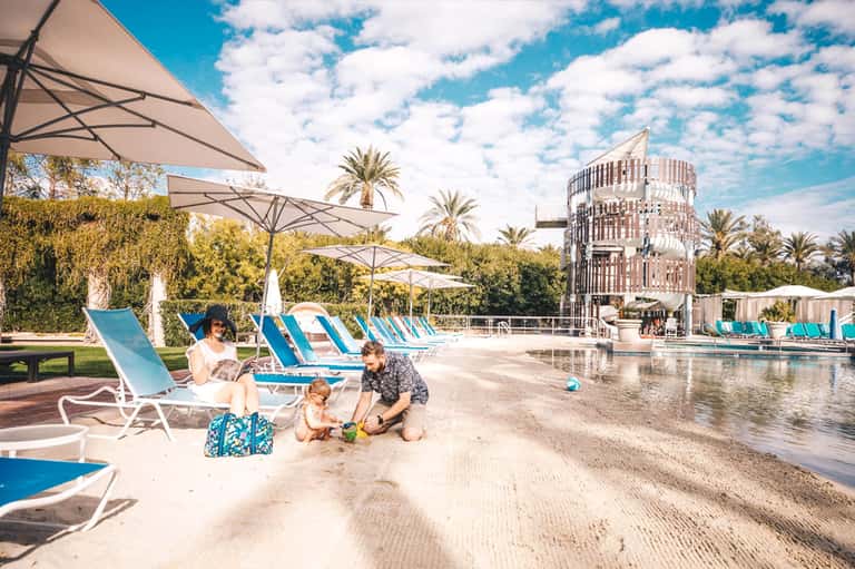 Family relaxing by a resort pool area with sandy beach, blue umbrellas, palm trees, and modern architecture