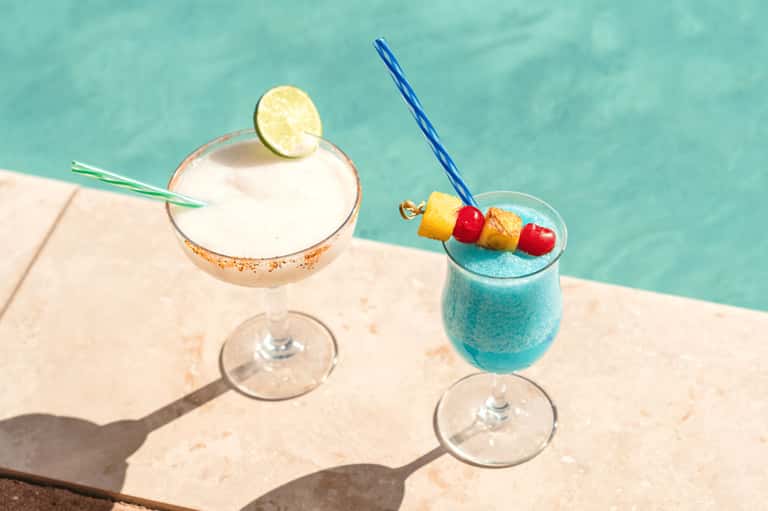Two colorful cocktails by turquoise pool with tropical garnishes and fruit skewers
