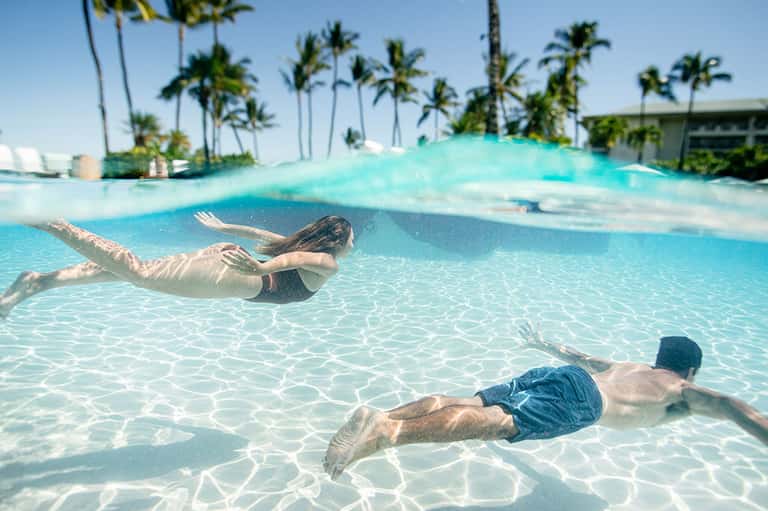Two people snorkeling in crystal-clear turquoise shallow water with palm trees visible