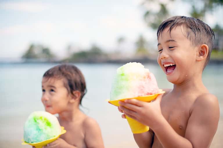 Two children enjoying colorful shaved ice treats on a tropical beach with palm trees