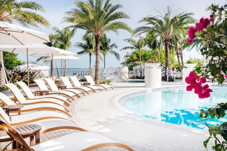 Luxury resort pool area with palm trees, white umbrellas, lounge chairs, and ocean view in background