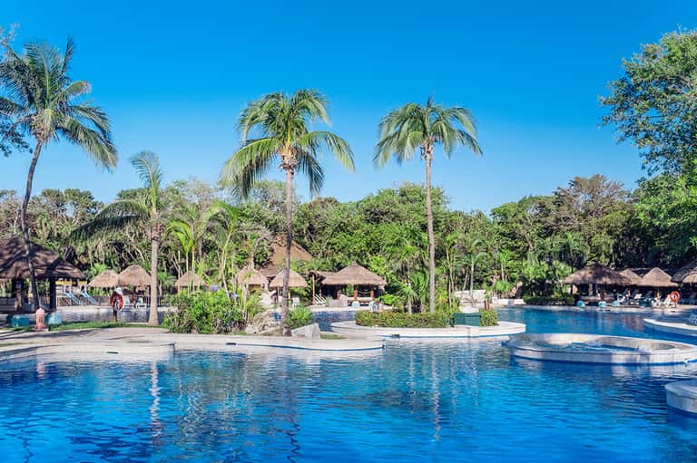 Resort pool with palm trees, thatched-roof cabanas, and tropical landscaping