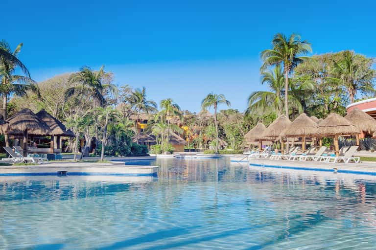 Resort pool area with thatched-roof palapas, palm trees, and lounge chairs under clear sky