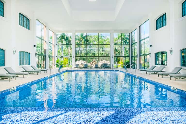 Indoor lap pool with high ceilings, large windows overlooking grounds, lounge chairs on sides