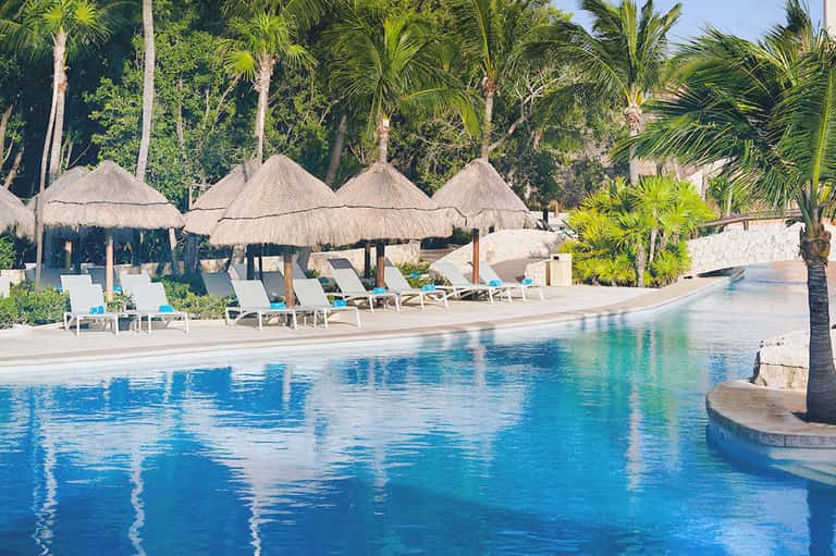 Resort pool with thatched umbrellas, white loungers, palm trees, and clear blue water
