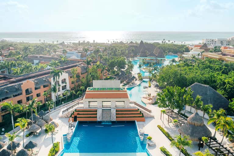 Aerial view of beachfront resort with pools, palm trees, ocean, and tiki-style structures