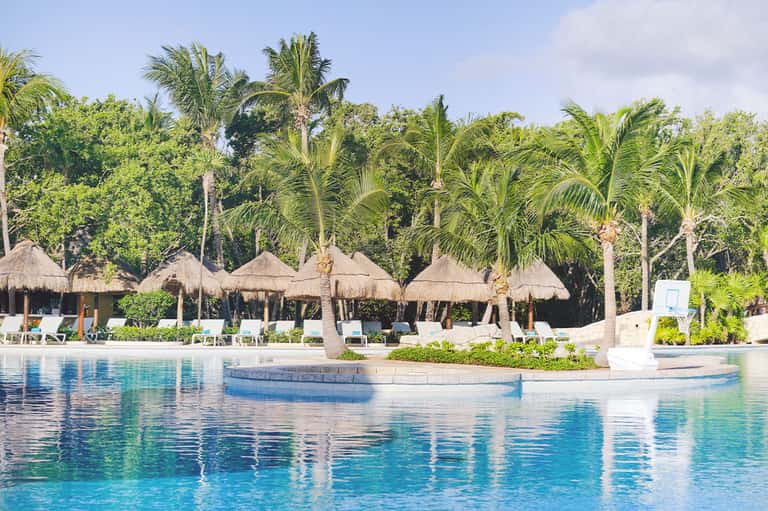Resort pool with thatched bungalows, palm trees, white loungers, and tropical island setting