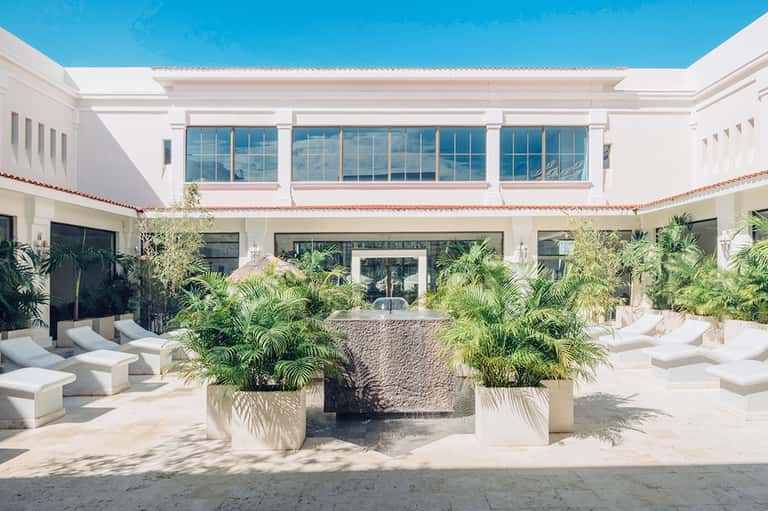 Modern resort courtyard with white lounge beds, tropical plants in planters, and white building with blue sky