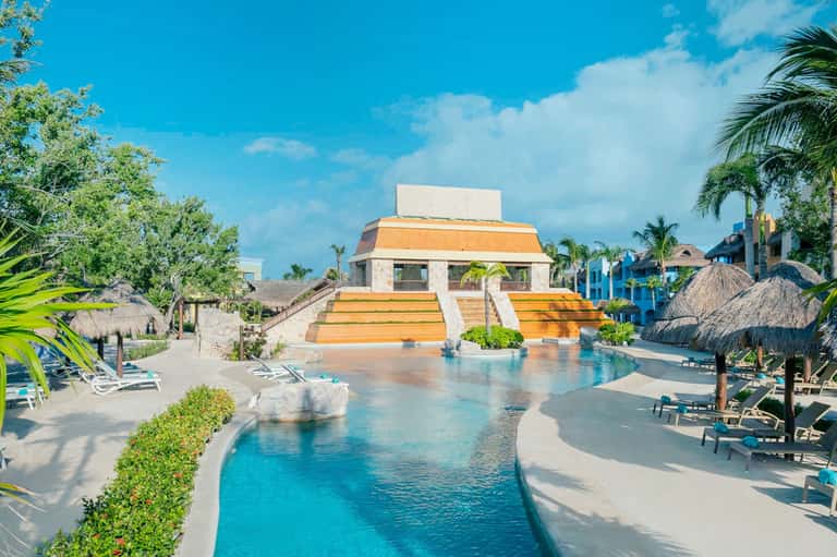 Luxury resort pool with Mayan pyramid-style architecture, palm trees, and lounge chairs surrounding crystal-clear water