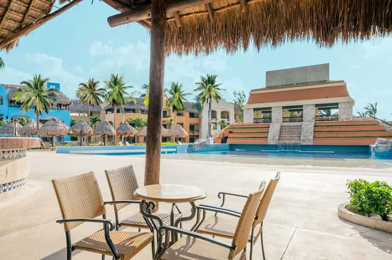 Beachfront resort terrace with thatched palapa, lounge chairs, pool, and multi-story hotel buildings