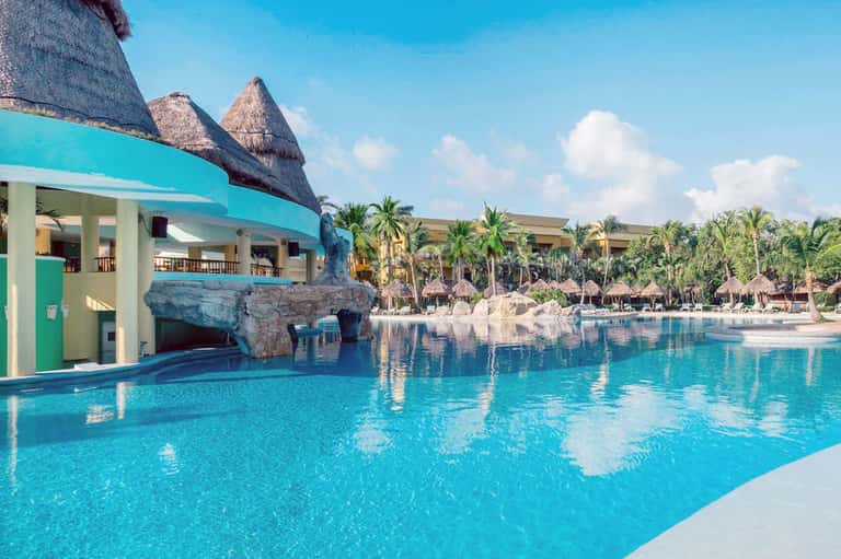 Resort swimming pool with turquoise water, palms, and thatched-roof structures