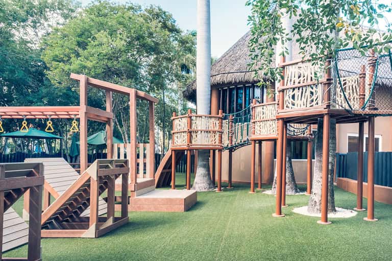 Wooden playground structure with climbing frames, slides, and thatched roof buildings in tropical setting