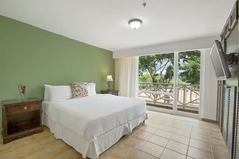 Guest bedroom with green accent wall, queen bed, white linens, balcony doors, and tree view