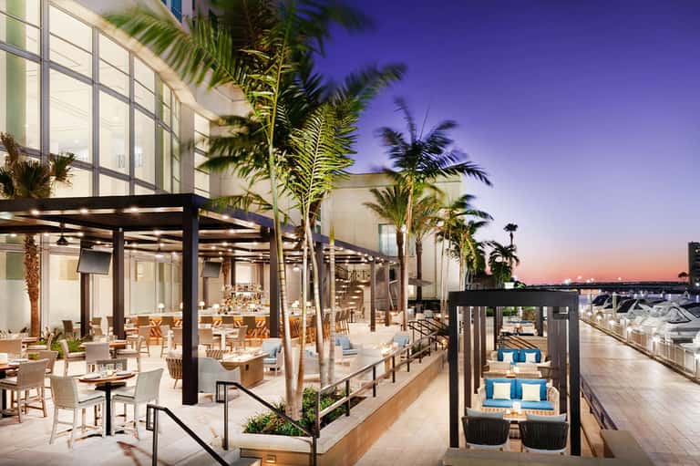 Modern waterfront restaurant terrace with dining tables, palm trees at dusk with marina views