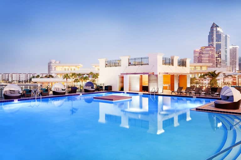 Resort pool at dusk with illuminated white cabanas and city skyline in background