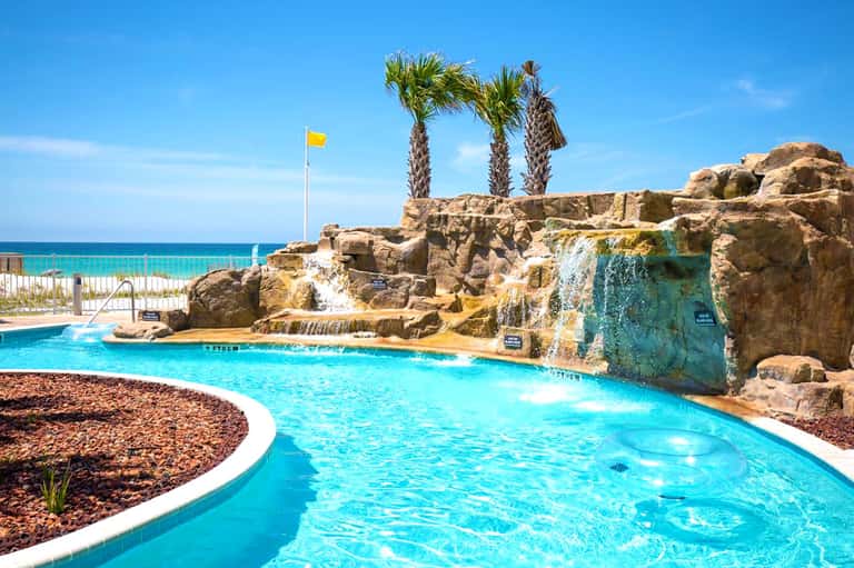 Turquoise resort pool with rocky features, palm trees, and ocean view in bright sunny weather