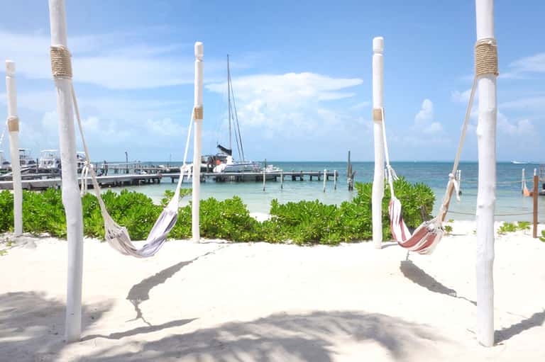White hammocks strung between poles on sandy beach with sailboat and dock