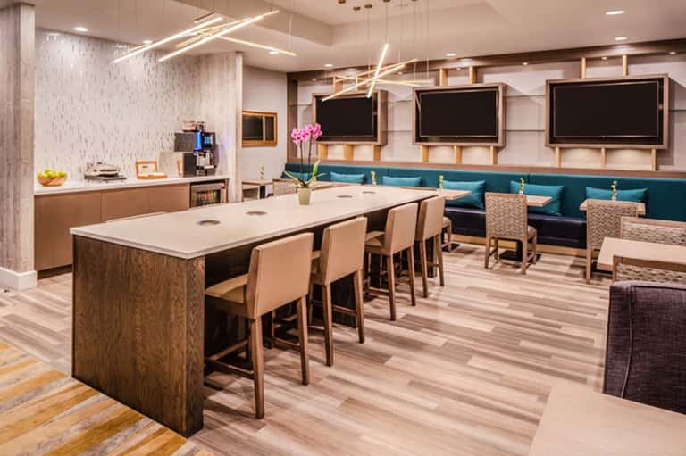 Modern hotel lounge with kitchen area, high-top table, seating, and flat-screen TVs