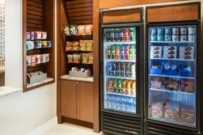 Hotel snack shop with wooden shelving displaying packaged snacks on left, refrigerated beverages and frozen items in glass cases on right