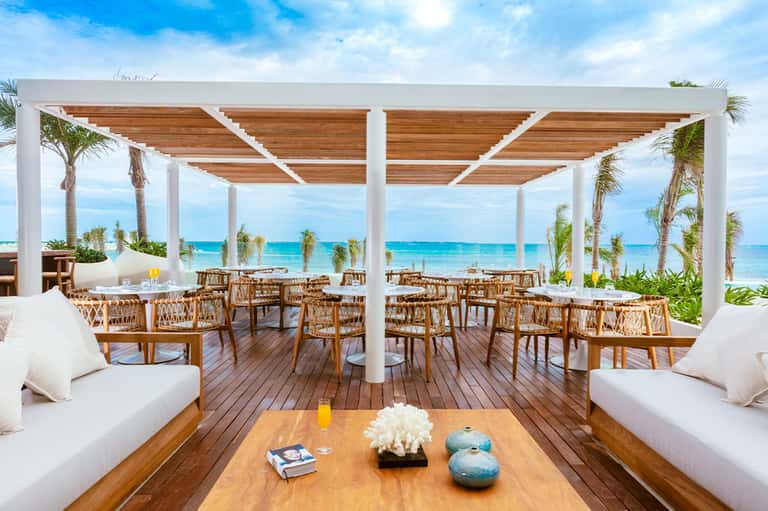 Beachfront dining pavilion with white pergola, wooden furniture, ocean views, and palm tree landscaping