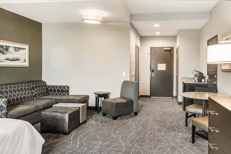 Hotel suite living room with patterned sofa, ottomans, kitchenette, and bathroom entrance