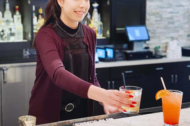 Female bartender in maroon shirt preparing cocktail at modern bar with bottles in background