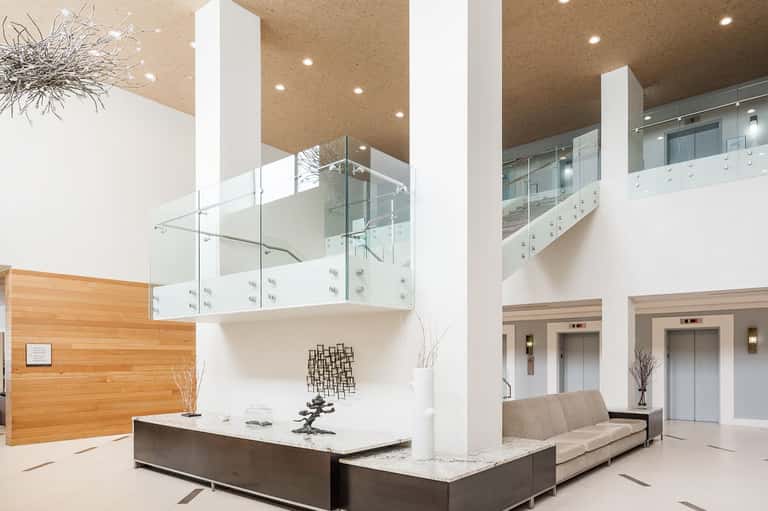 Modern hotel atrium with multi-level glass railings, white columns, reception desk, and brass ceiling