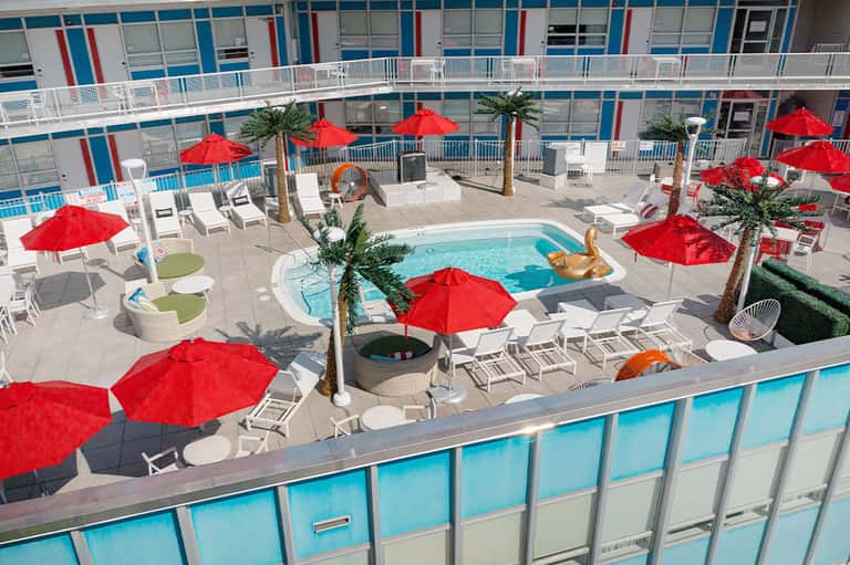 Rooftop pool deck with bright red umbrellas, white lounge chairs, palm trees, and colorful building facade