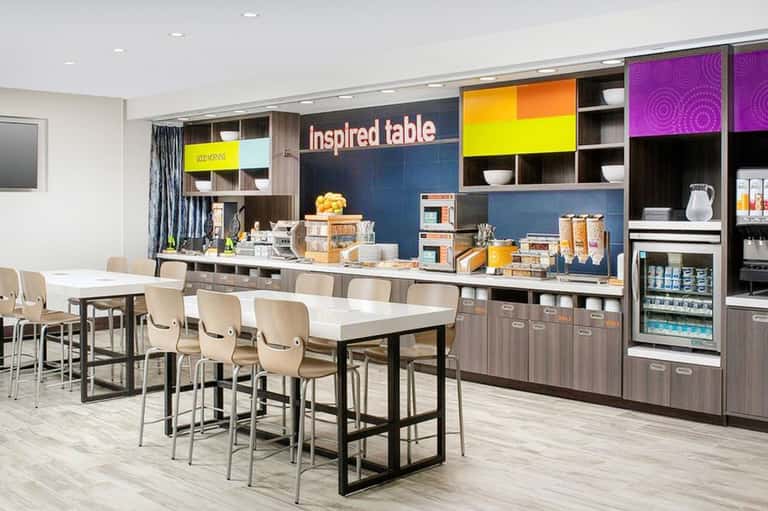 Modern hotel break room with colorful shelving, kitchenette, high-top dining table, and beige chairs