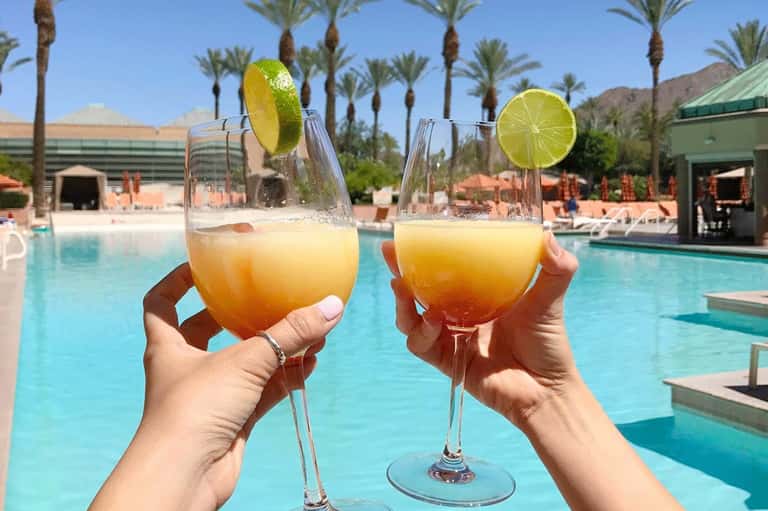 Two tropical cocktails with lime slices by a resort pool surrounded by palm trees and desert mountains