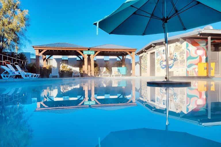 Modern pool with wooden pergola, blue umbrella, and reflected sky creating mirror-like surface