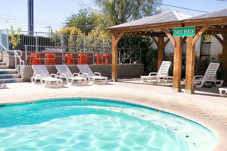 Resort pool with wooden pergola, white lounge chairs, and Oasis Beach sign
