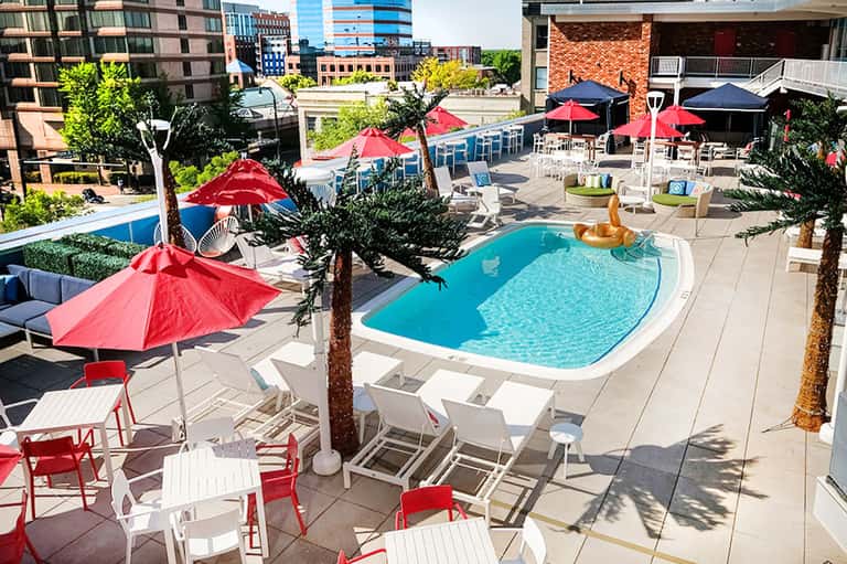 Urban rooftop pool with red umbrellas, palm trees, and modern high-rise buildings in background