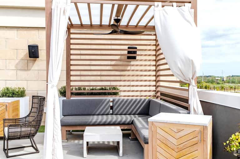 Modern outdoor living space with gray sectional sofa under wooden pergola with white curtains and ceiling fan