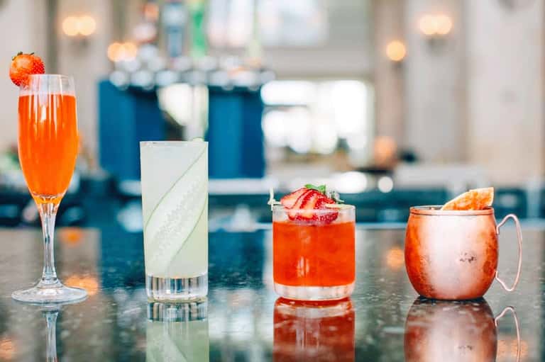 Four colorful cocktails on a bar counter with orange, white, and red beverages garnished with fruit