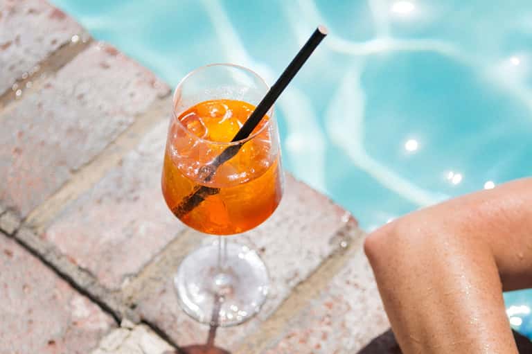 Orange cocktail with ice and orange slice on poolside brick deck with turquoise water background