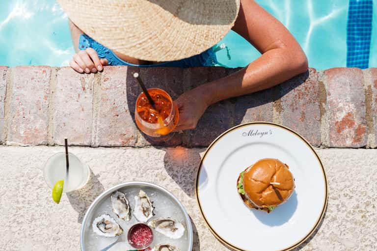 Person in sun hat poolside holding tropical cocktail with oysters and burger plates nearby