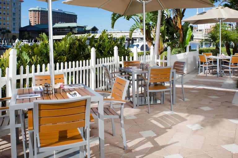 Outdoor beachfront dining area with white picket fencing, umbrellas, and wooden chairs overlooking the water