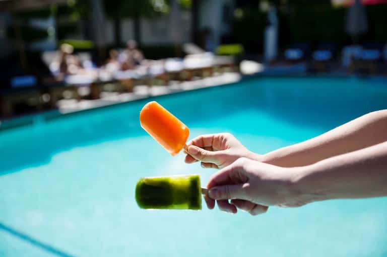 Hands holding colorful popsicles over a bright turquoise swimming pool