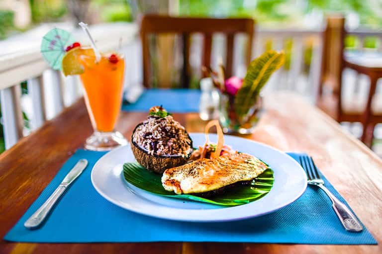Beachfront dining table with grilled fish, stuffed coconut, tropical orange cocktail, and water view